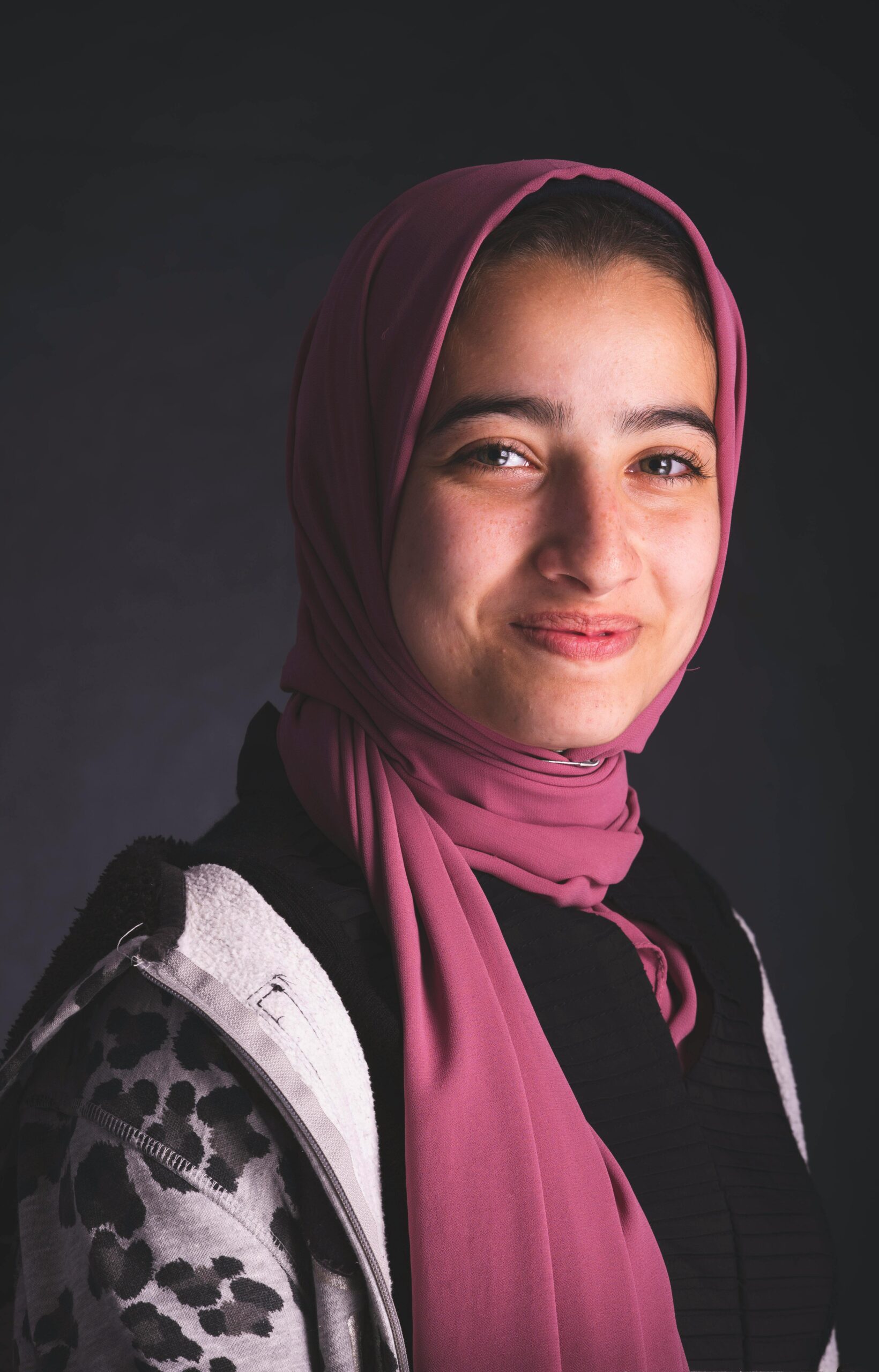 A thoughtful portrait of a smiling young woman wearing a pink hijab against a dark background.